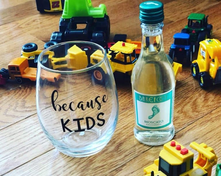 Stemless wine glass with black Cricut vinyl lettering reading 'because KIDS' next to a mini bottle of Barefoot Moscato, surrounded by scattered children's toy trucks on a wooden floor