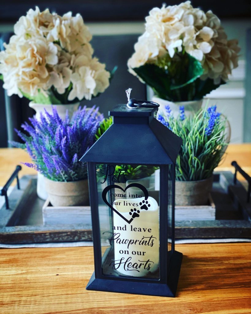 How to Price Your Cricut Crafts: A Detailed Guide 1 A black lantern with decorative cricut permanent vinyl text and memorial symbols on a wooden table, surrounded by potted flowers including white hydrangeas and purple lavender, creating a cozy home decor scene.