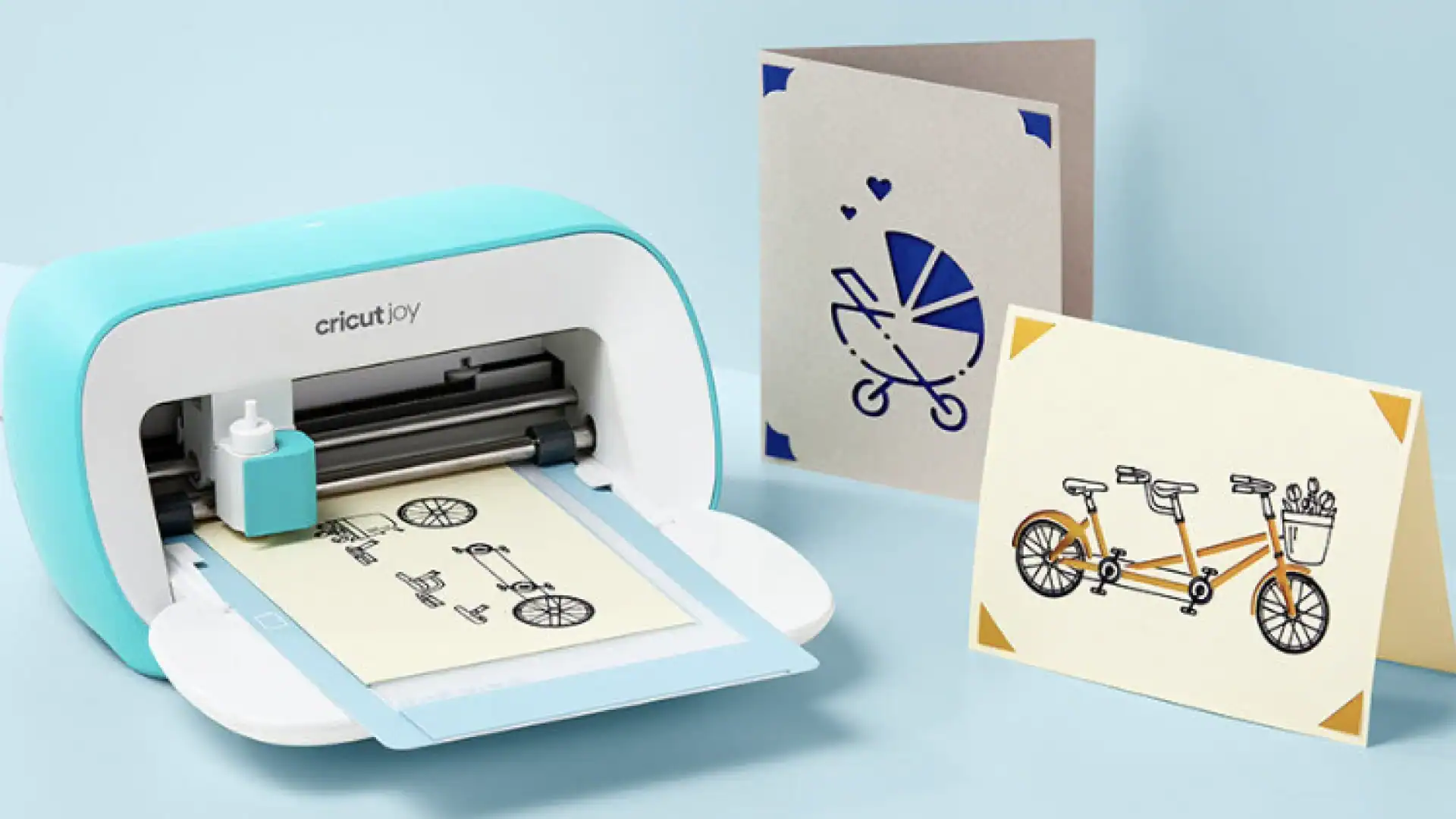 Teal and white Cricut Joy compact cutting machine drawing a tandem bicycle design on cream-colored card stock using the pen feature. Two finished handmade greeting cards are displayed beside it: a gray card with blue baby stroller design and hearts, and a cream card with an orange tandem bicycle carrying a basket of flowers. Both cards feature decorative corner accents. Light blue background showcases the machine's card-making capabilities.