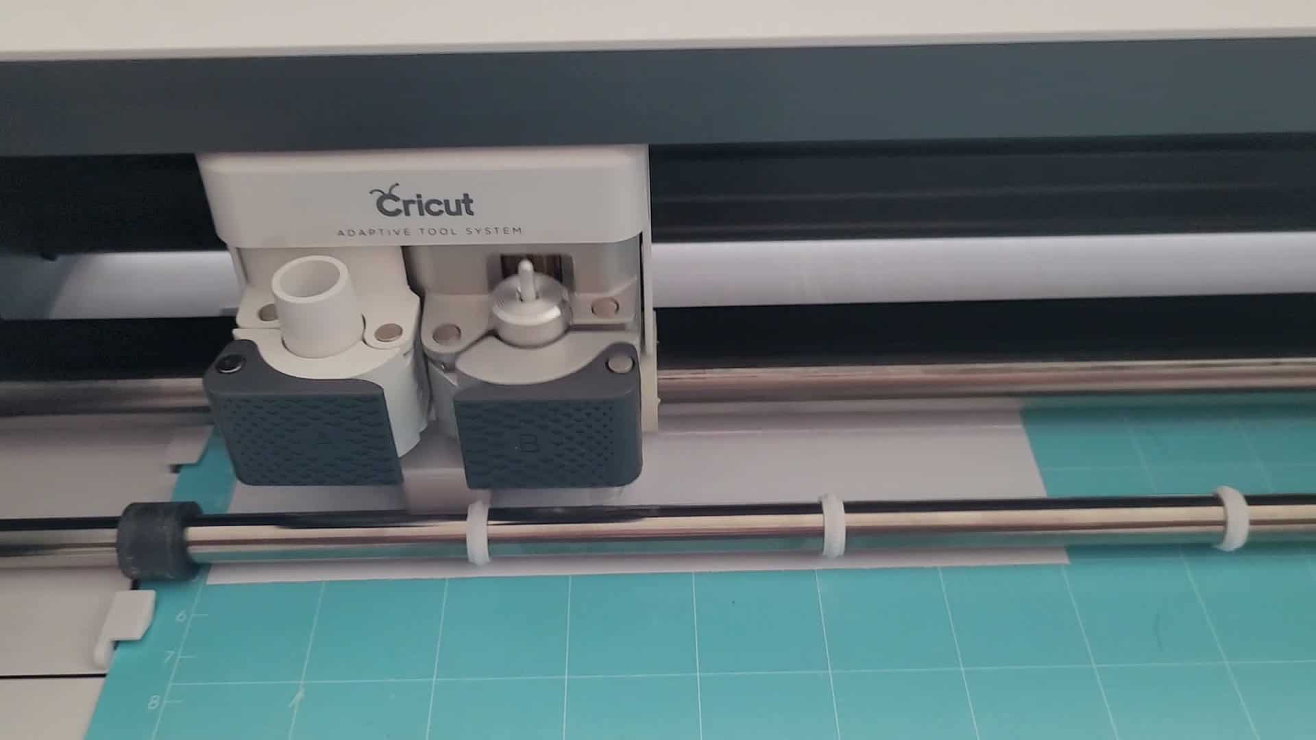 Close-up view of the Cricut Adaptive Tool System carriage on a Cricut Maker machine, showing the dual tool clamps labeled A and B with textured gray grips. Clamp A is empty and ready for a tool, while clamp B contains the blade housing with adjustment gear. A teal StandardGrip cutting mat is loaded beneath with gray vinyl material positioned for cutting.