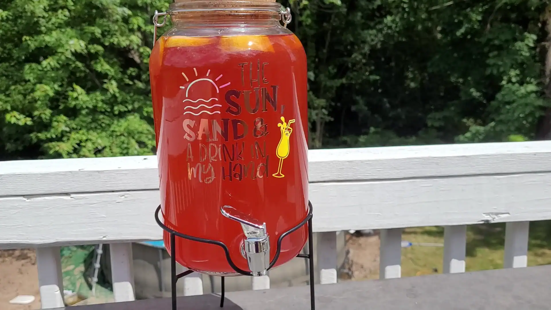 Glass beverage dispenser with cork lid filled with red-orange punch, decorated with colorful adhesive vinyl reading "The Sun, Sand & A Drink in My Hand" alongside a sun and waves graphic and a yellow tropical cocktail illustration. The dispenser sits in a black metal stand with chrome spigot on an outdoor deck, with green summer trees in the background.