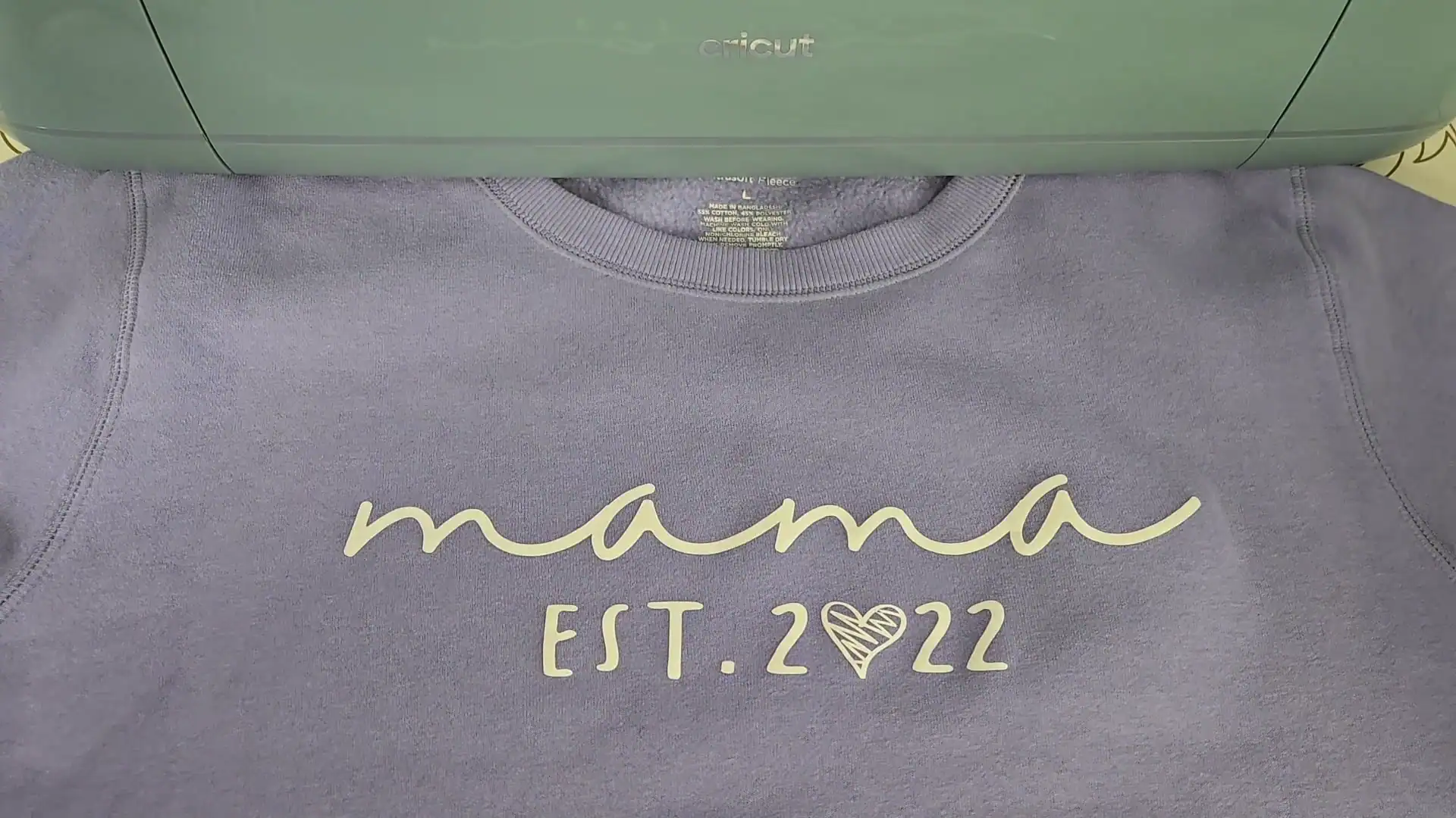 Gray crewneck sweatshirt customized with light yellow heat transfer vinyl featuring "mama" in flowing cursive script and "EST. 2022" below it, with a decorative scribble heart replacing the zero. A sage green Cricut machine is visible at the top of the frame, showing the tool used to cut the design.