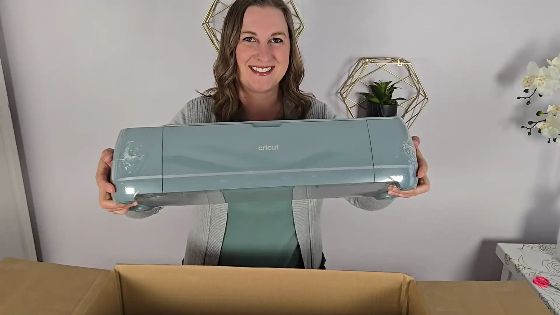 Kerri Adamczyk smiling while holding up a new sage green Cricut Maker 4 machine fresh from unboxing, with protective plastic wrap still on the ends. A cardboard shipping box is visible in the foreground, and the background features gold geometric wall decor with a small plant.