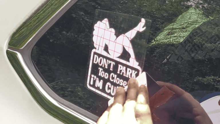Kerri Adamczyk applying a custom vinyl car decal with the text 'Don't Park Too Close I'm Cute' onto a car window. The decal features a silhouette design and transfer tape is visible. Ideal example for a DIY guide using Cricut 2025.
