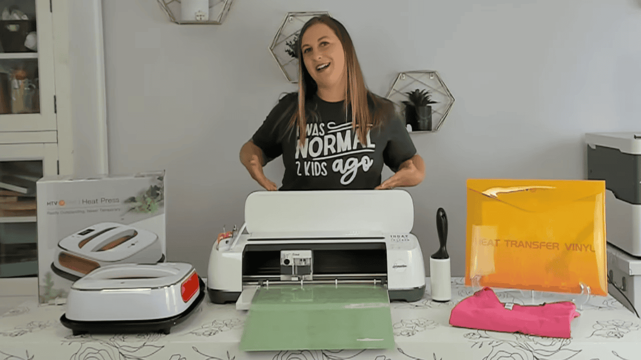 Kerri smiling behind a Cricut Maker with an HTVRont heat press, orange heat transfer vinyl and a pink t-shirt