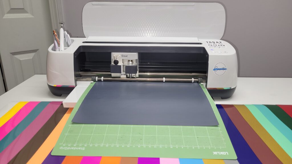 Cricut cutting machine with a gray cutting mat and various colorful vinyl sheets arranged nearby.