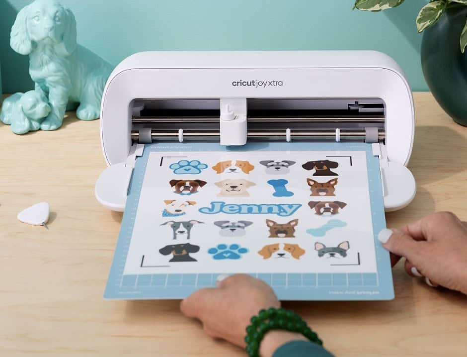 Cricut Joy Xtra machine cutting a Print Then Cut sticker sheet featuring cute dog breed faces, blue paw prints, bone shapes, and the name 'Jenny' on a light blue cutting mat, with hands guiding the mat on a wooden desk with teal dog figurine and plant in background