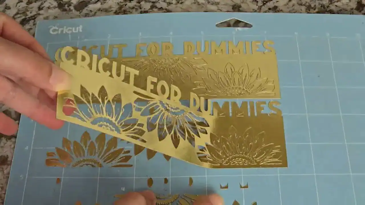 Hand carefully lifting delicate gold foil cardstock bookmark with intricate floral cutouts from blue LightGrip Cricut mat