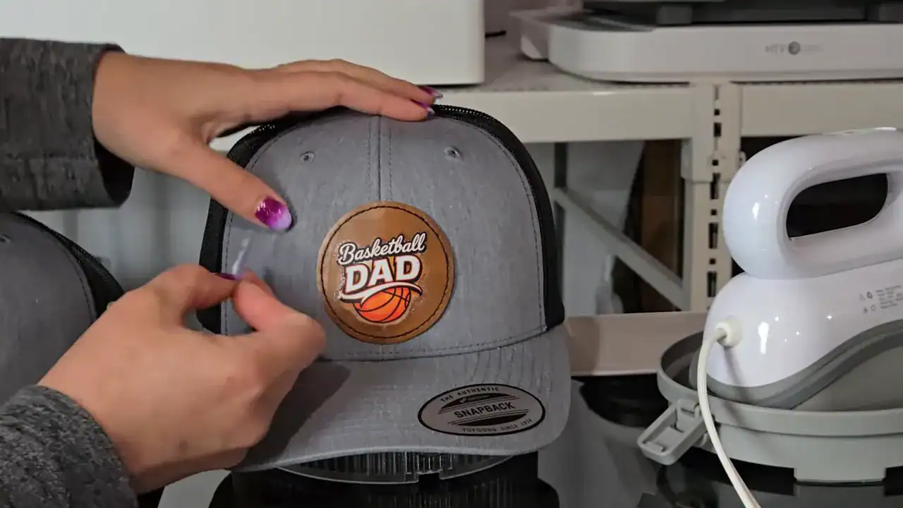 Hands holding a gray trucker hat with a faux leather patch reading ‘Basketball Dad,’ DTF printed using the xTool Apparel Printer and pressed with a mini heat press.