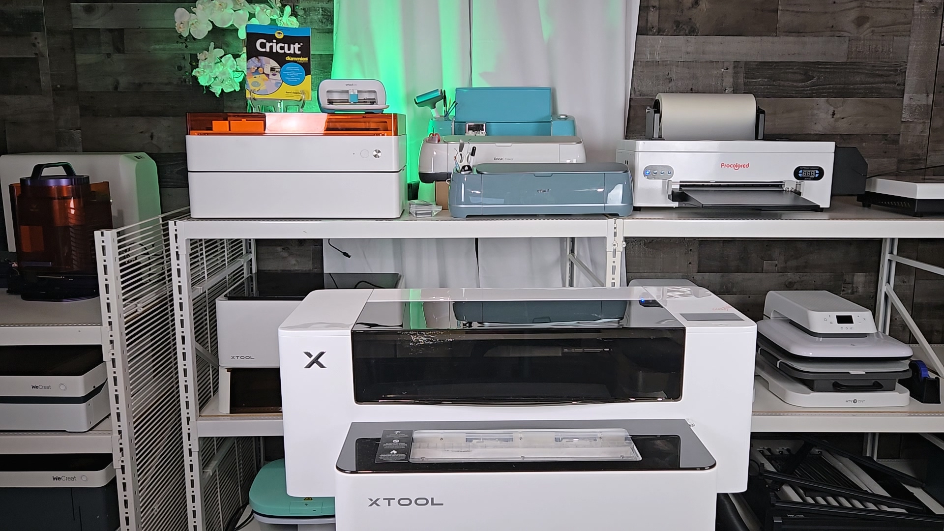 Craft room workspace showing xTool Apparel Printer stacked on shaker oven in center, with Procolored F13 Panda DTF printer on shelf to the right, surrounded by various crafting machines including Cricut and laser engravers