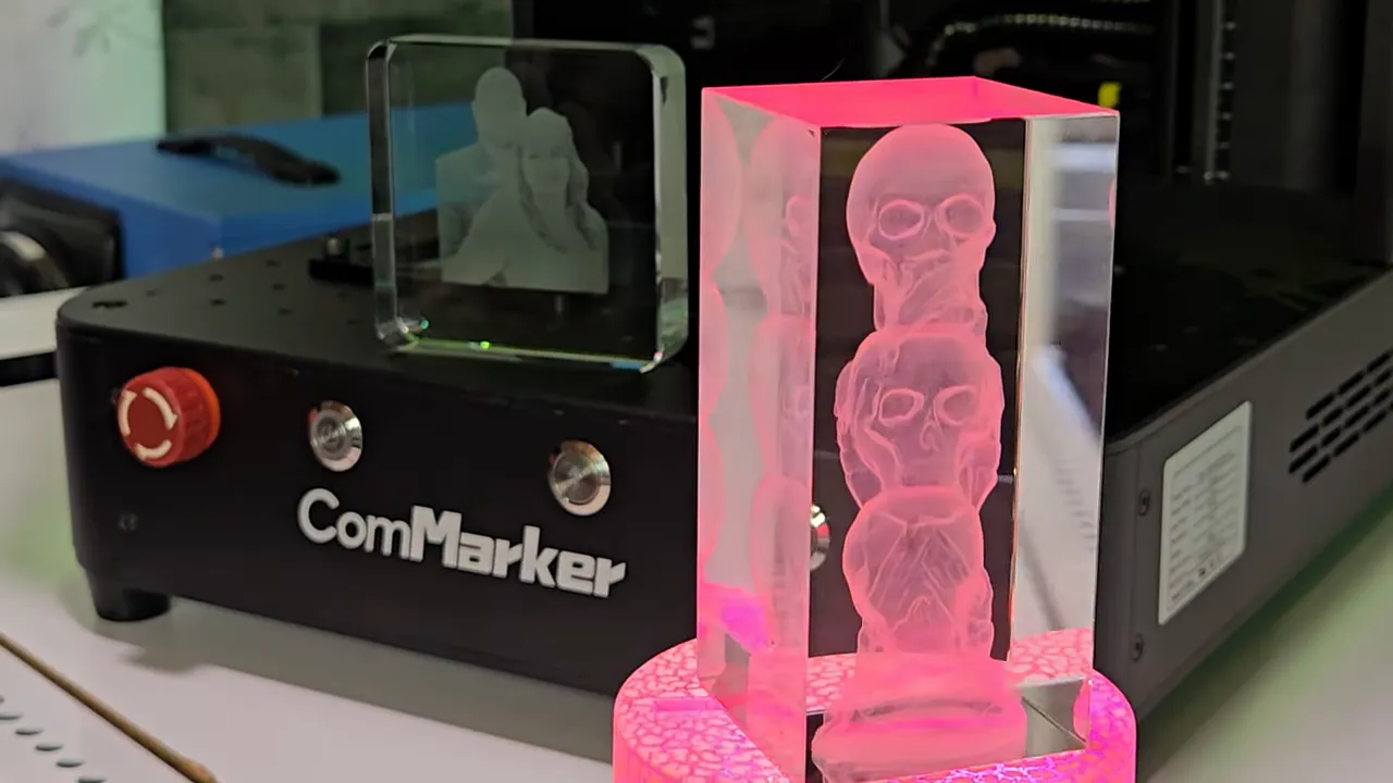 ComMarker Omni X Review: The New Way To Do 3D Glass Engraving at Home 10 Alt Text: Two 3D laser-engraved crystal pieces created with the ComMarker Omni X displayed in front of the machine. The tall rectangular crystal features a stacked skulls design illuminated by a pink LED light base with a crackle pattern, while a smaller square crystal with a 2D couple's photo engraving sits on top of the ComMarker unit. The machine's black control panel with red emergency stop button and silver knobs is visible, along with a blue storage case in the background.