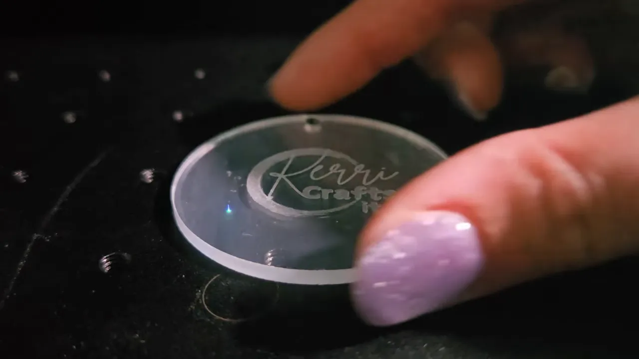 ComMarker Omni X Review: The New Way To Do 3D Glass Engraving at Home 9 Kerri Adamczyk with sparkly lavender nail polish positioning a round clear acrylic disc with the "Kerri Crafts It" logo laser-engraved on it. The acrylic keychain blank sits on the perforated metal work bed of a laser engraver, with a small blue laser dot visible on the surface during the engraving process.