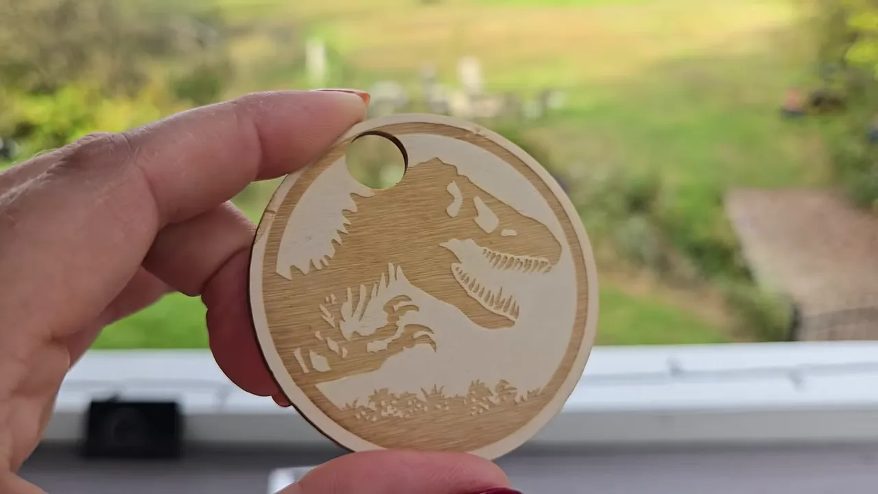 ComMarker Omni X Review: The New Way To Do 3D Glass Engraving at Home 8 Kerri Adamczyk holding a round laser-engraved wooden ornament featuring a Jurassic Park-style T-Rex dinosaur skeleton silhouette design with tropical foliage accents. The ornament has a hole at the top for hanging and shows contrasting light and dark wood tones from the engraving process. A blurred backyard scene with green grass is visible through a window in the background.