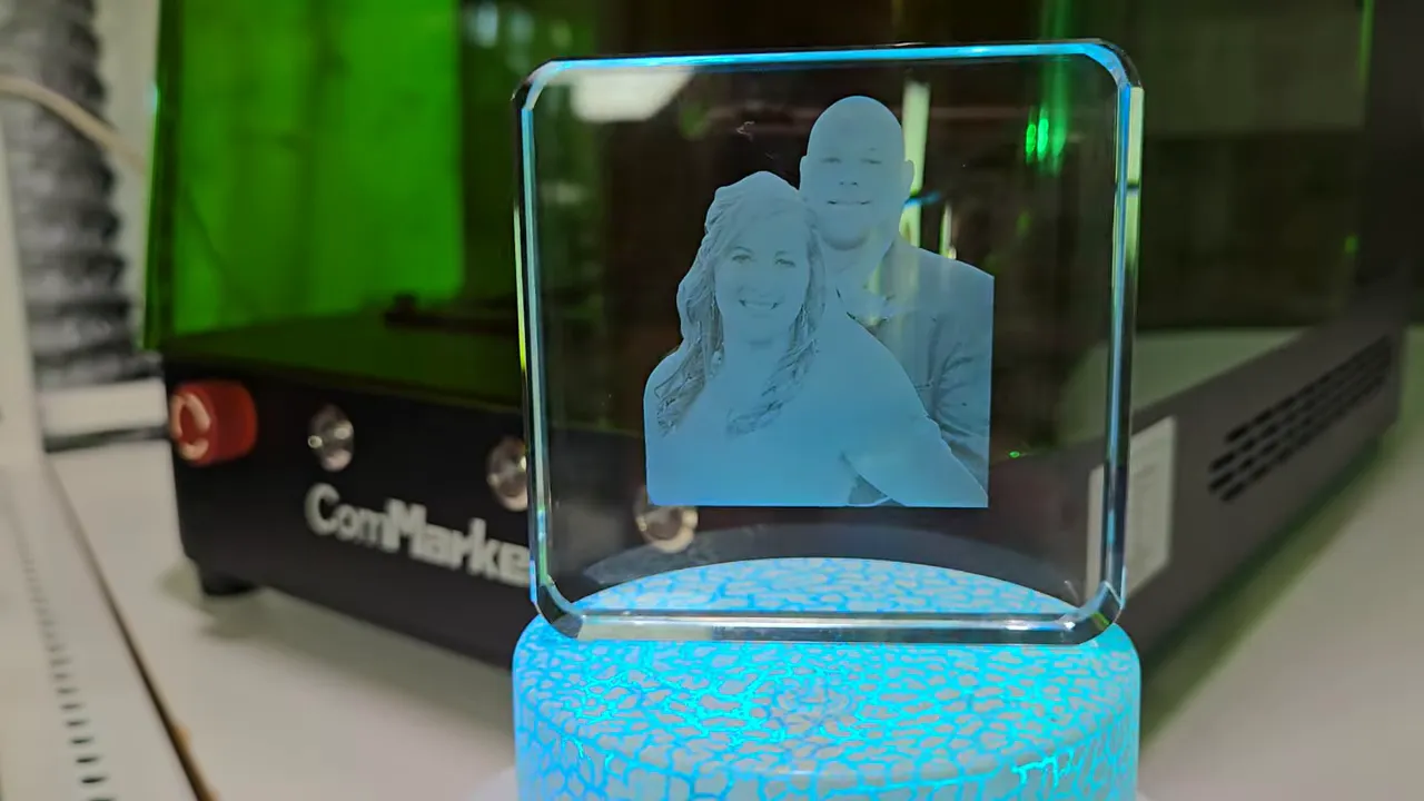 ComMarker Omni X Review: The New Way To Do 3D Glass Engraving at Home 11 2D laser-engraved crystal photo of a smiling couple displayed on a blue LED light base with a crackle pattern, created using the ComMarker Omni X. The square crystal block sits in front of the ComMarker machine with its green-tinted safety enclosure visible in the background.