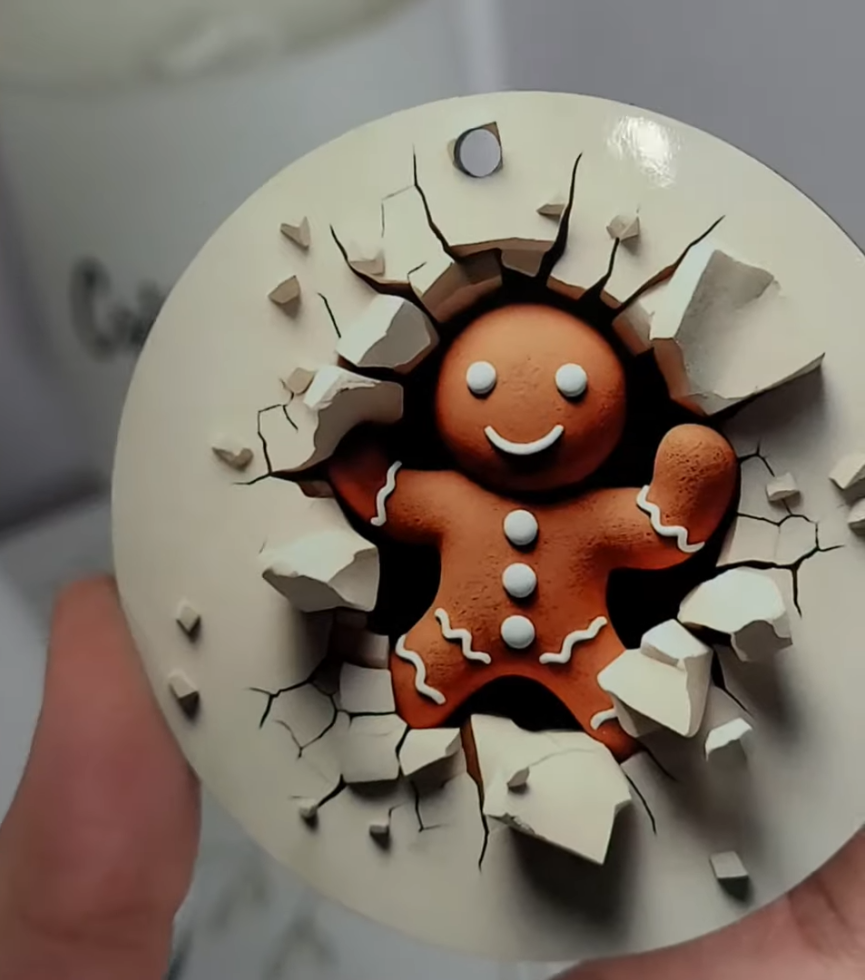 How to Make 3D Sublimation Ornaments That Pop Right Off the Surface 1 kerri adamczyk holding a round sublimated Christmas ornament featuring a 3D optical illusion design of a smiling gingerbread man appearing to burst through a cracked white wall. The gingerbread man has white icing details on its arms and legs, white button decorations, and a cheerful smile. The design creates a realistic depth effect with broken wall pieces and cracks surrounding the character. A small hole at the top of the ornament allows for hanging.