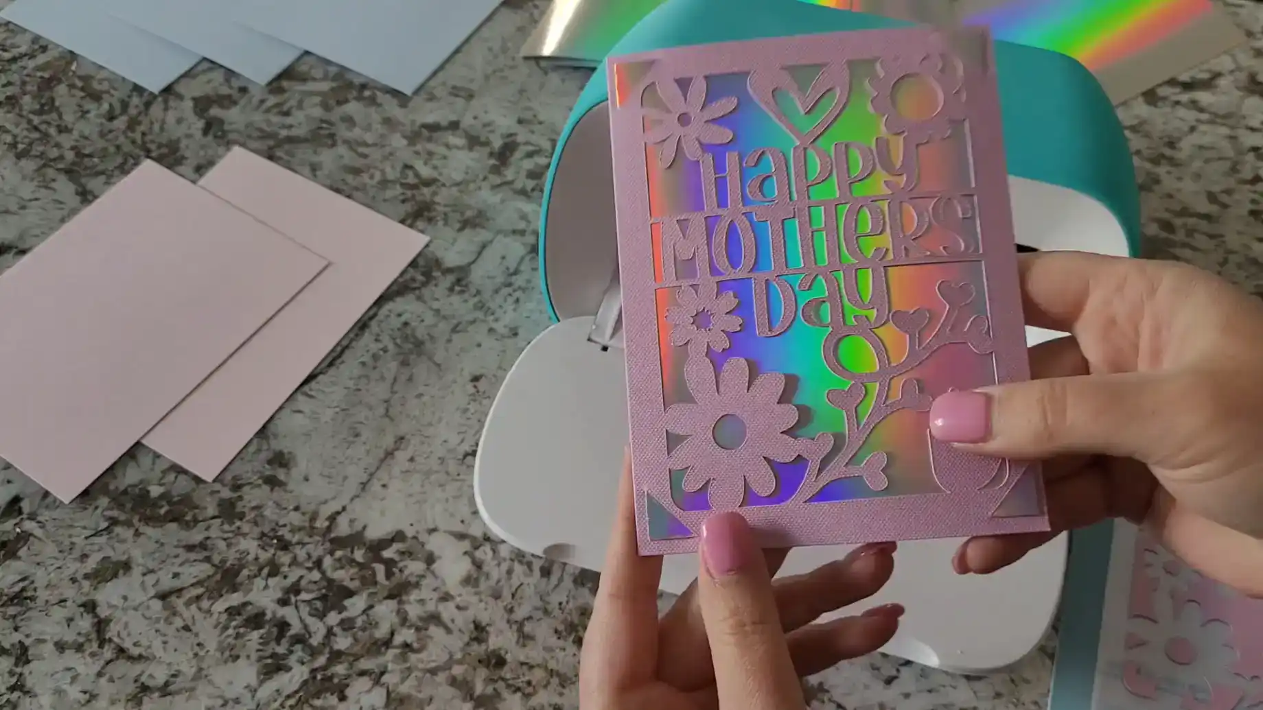 Kerri Adamczyk with pink nail polish holding a handmade Happy Mother's Day card with intricate floral cutout design in pink cardstock over holographic rainbow insert, made with Cricut Joy visible in background alongside card blanks and envelopes on granite countertop