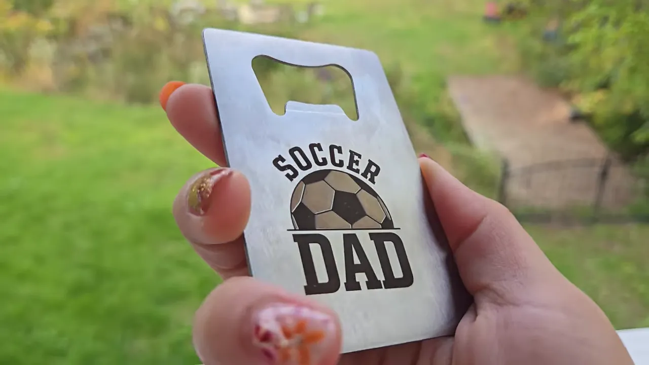 ComMarker Omni X Review: The New Way To Do 3D Glass Engraving at Home 7 Kerri Adamczyk with colorful nail art holding a stainless steel credit card-style bottle opener featuring a laser-engraved "SOCCER DAD" design with a soccer ball graphic, created using the ComMarker Omni X by combining light and dark engraving settings. The bottle opener cutout is visible at the top of the card, with a blurred backyard scene showing green grass and a stone pathway in the background.