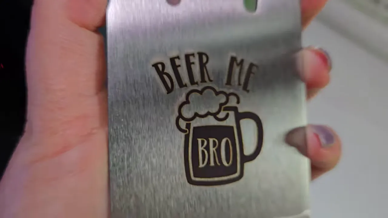 ComMarker Omni X Review: The New Way To Do 3D Glass Engraving at Home 6 Kerri Adamczyk Alt Text: Hands holding a brushed stainless steel credit card-style bottle opener with a dark laser-engraved design featuring "BEER ME" text above a foaming beer mug with "BRO" written on it. This version shows a darker, more contrasted engraving compared to a lighter etching style, with the bottle opener notch partially visible at the top edge.