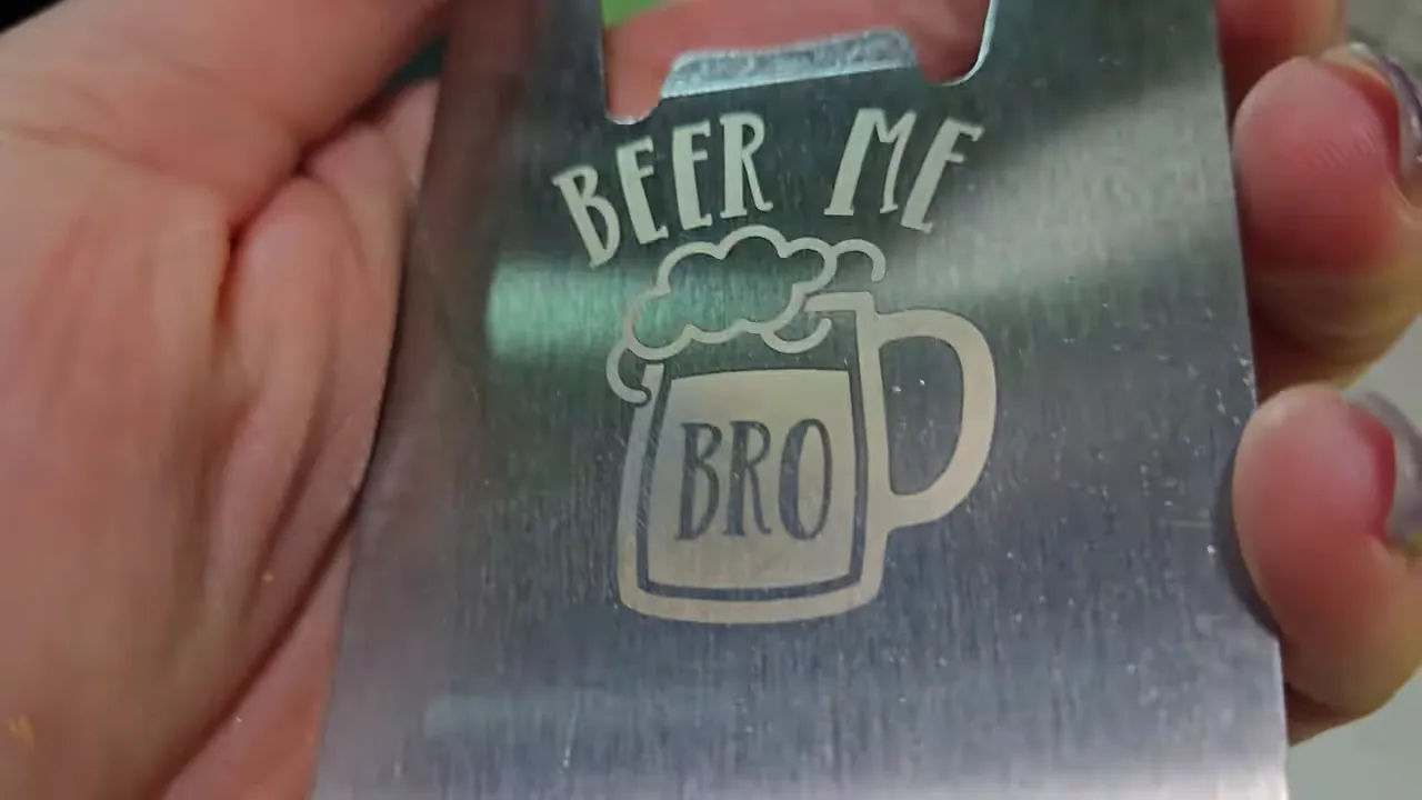 ComMarker Omni X Review: The New Way To Do 3D Glass Engraving at Home 5 Kerri Adamczyk holding a brushed stainless steel credit card-style bottle opener with a laser-engraved design featuring the text "BEER ME" above a foaming beer mug graphic with "BRO" written on the mug. The bottle opener notch is visible at the top edge of the metal card.