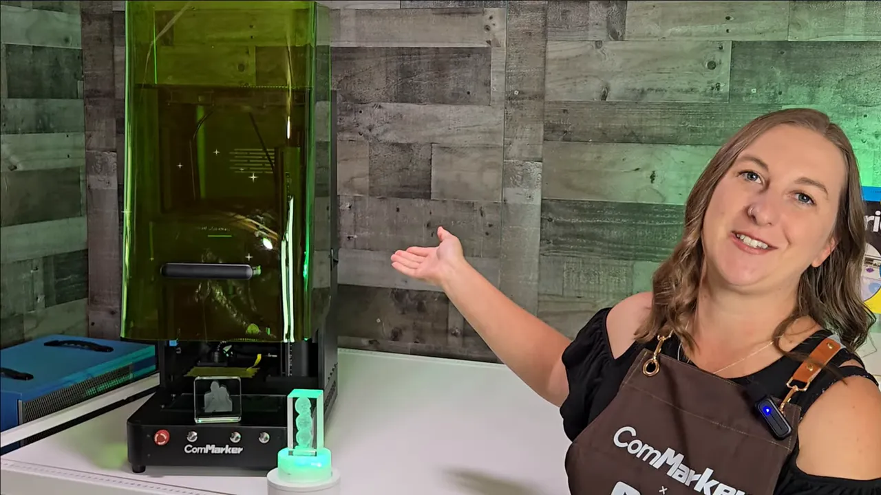 ComMarker Omni X Review: The New Way To Do 3D Glass Engraving at Home 1 Kerri Adamczyk wearing a black top and brown ComMarker-branded apron gestures toward a ComMarker Omni X 3D crystal laser engraving machine with a tall green-tinted safety enclosure. On the white table in front of the machine sit two 3D laser-engraved crystal pieces, one displayed on a color-changing LED light base glowing green. A rustic gray wood plank wall serves as the backdrop, with green accent lighting visible on the right side.
