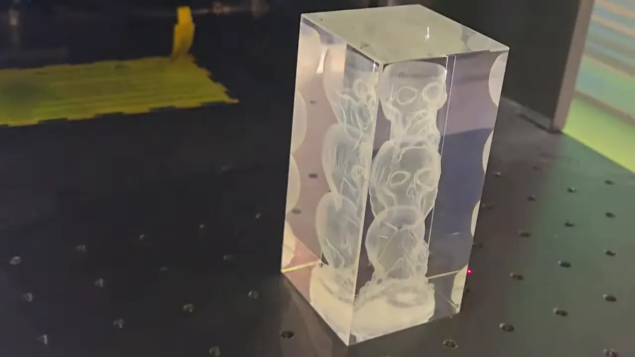 ComMarker Omni X Review: The New Way To Do 3D Glass Engraving at Home 2 3D laser-engraved crystal block featuring a stacked skulls design viewed from an angle, created using the ComMarker Omni X. The tall rectangular crystal sits on the machine's perforated metal work bed, with a pink laser alignment dot visible near the base and yellow positioning markers in the background. Colorful light reflections are visible on the right side of the frame.