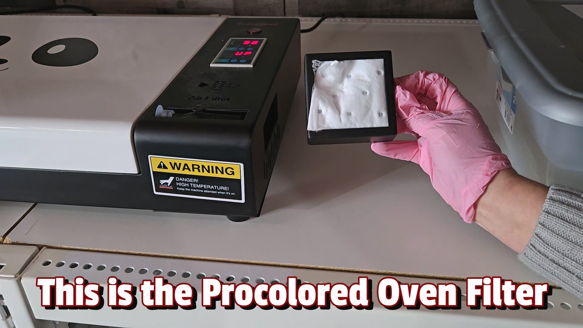 Kerri Adamczyk's Gloved hand holding Procolored oven filter tray showing white filter material, with smokeless oven unit displaying warning label about high temperature in background