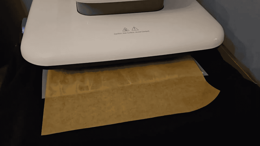 A heat press in the closed/pressing position during the DTF transfer application process. The white upper platen displays caution symbols and text reading "Caution: Hot Surface. Avoid Contact." Brown kraft/parchment protective paper covers the transfer area, acting as a barrier between the hot platen and the DTF transfer to prevent scorching or the transfer sticking to the press. The dark sweatshirt with the MAMA transfer is underneath, visible only at the edges. Using protective paper or a Teflon sheet is a standard practice when heat pressing transfers to protect both the design and the heat press surface. The press is applying the 320°F heat and 170 LB pressure for the 15-second duration shown in the previous settings image.
