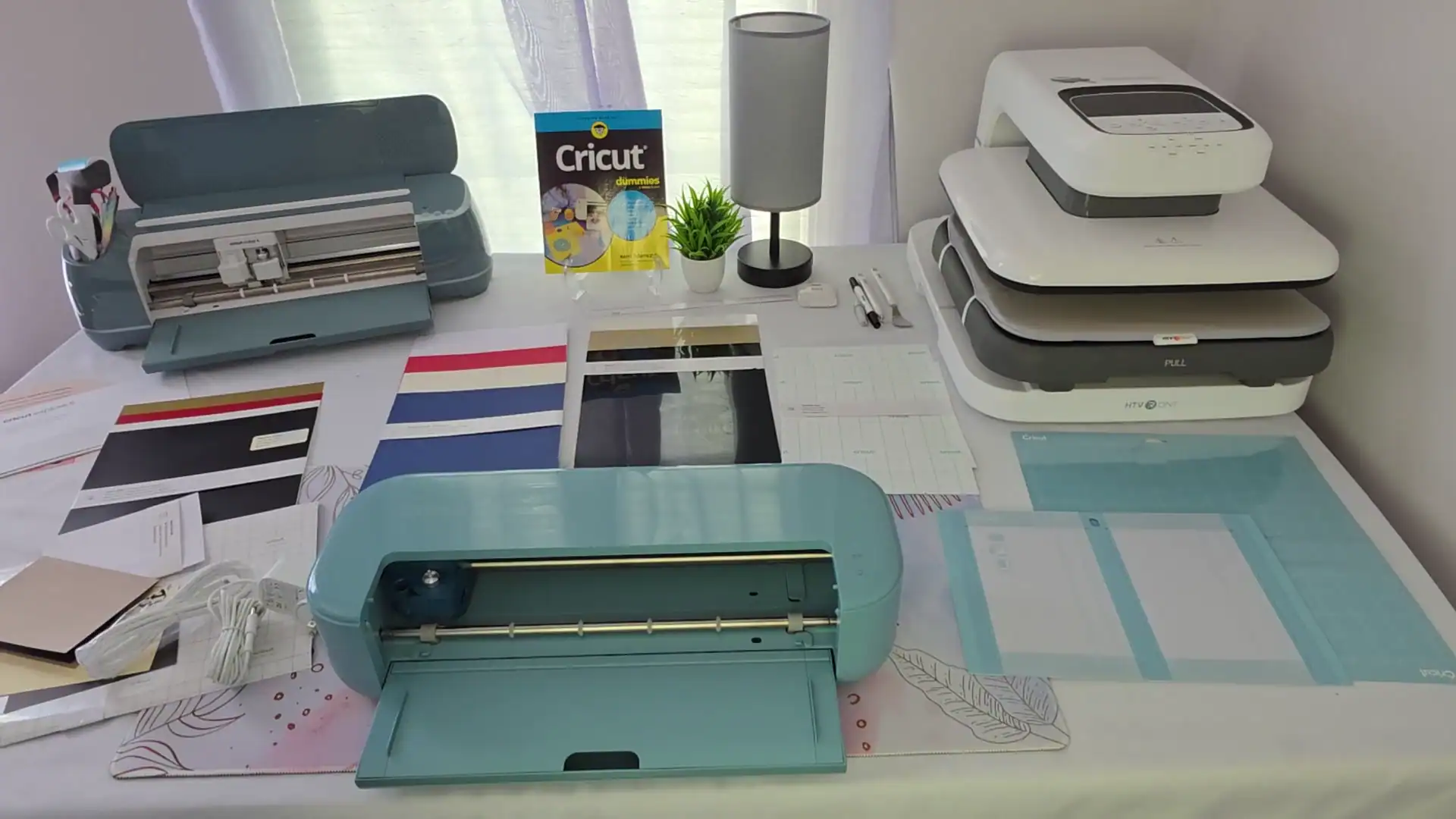 Cricut Explore 5 cutting machine unboxed on a crafting desk surrounded by vinyl sheets, cutting mats, accessories, a Cricut Maker 4, HTV Ont heat press, and Cricut For Dummies book.