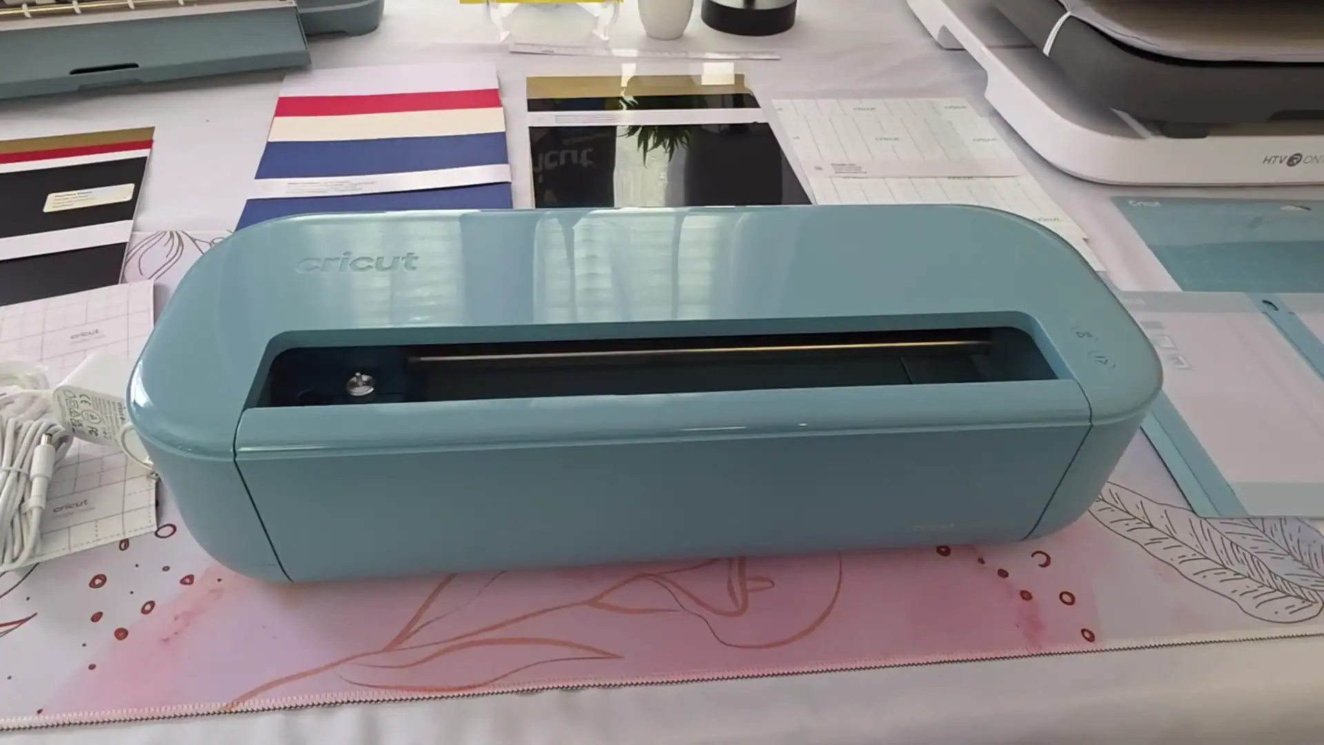 Close-up of the Cricut Explore 5 in teal showing its open-top carriage design with the cutting bar fully exposed, sitting on a crafting desk with vinyl sheets and cutting mats.