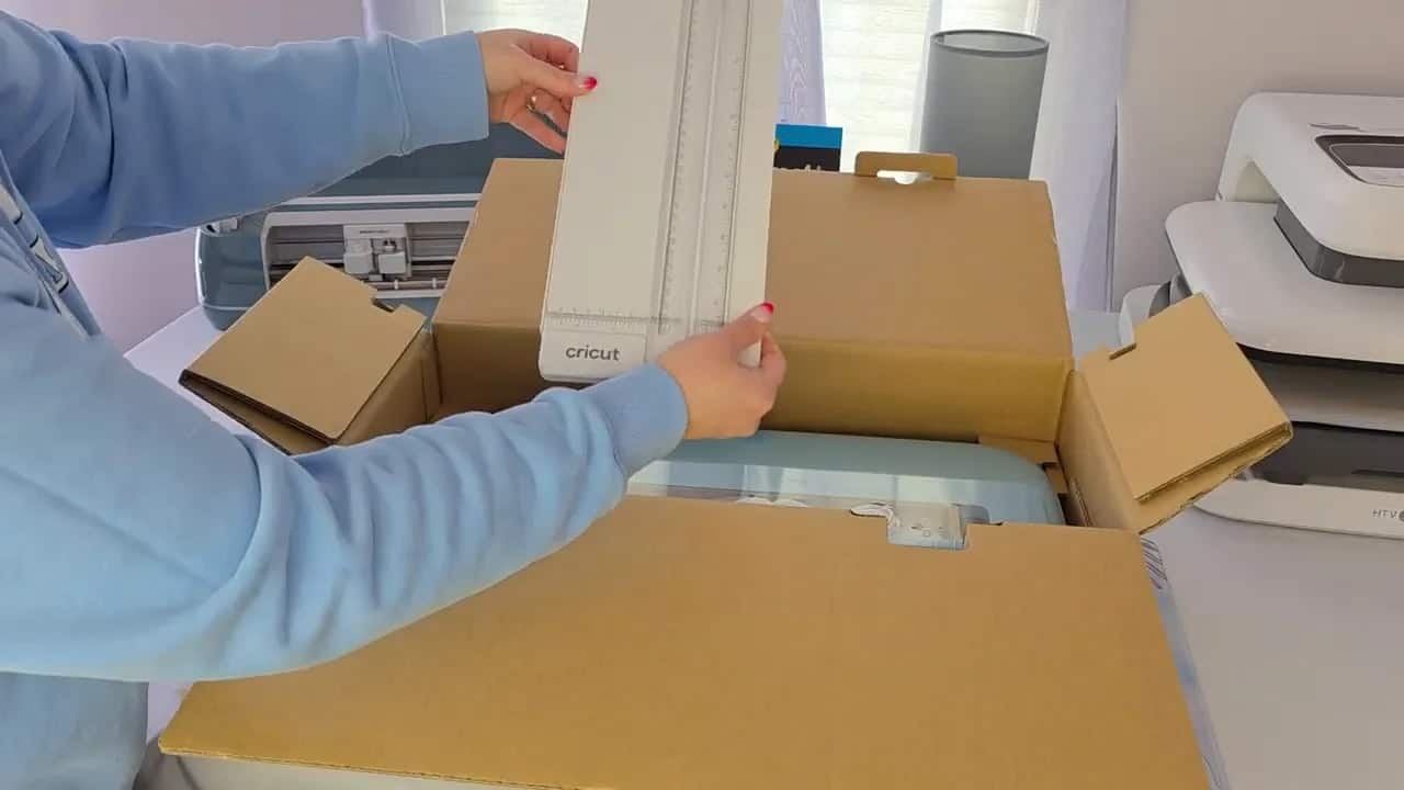 Teal Cricut Explore 5 partially visible in its box with the included ruler being lifted out.