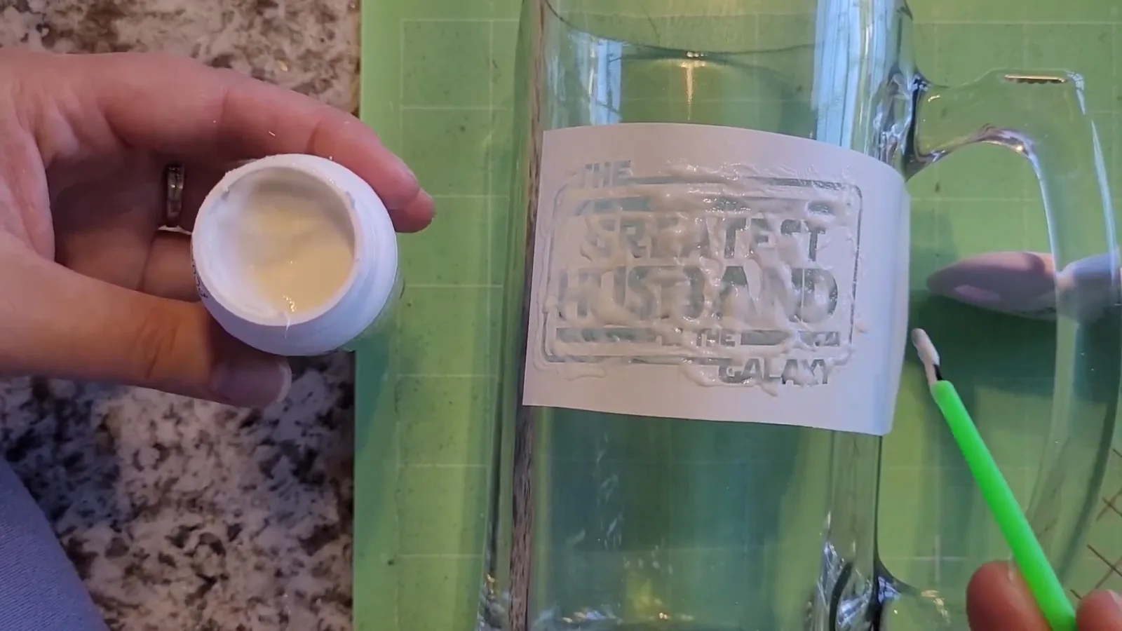 Hand holding an open jar of Armour Etch cream while a green paintbrush spreads it over the vinyl stencil on a glass mug
