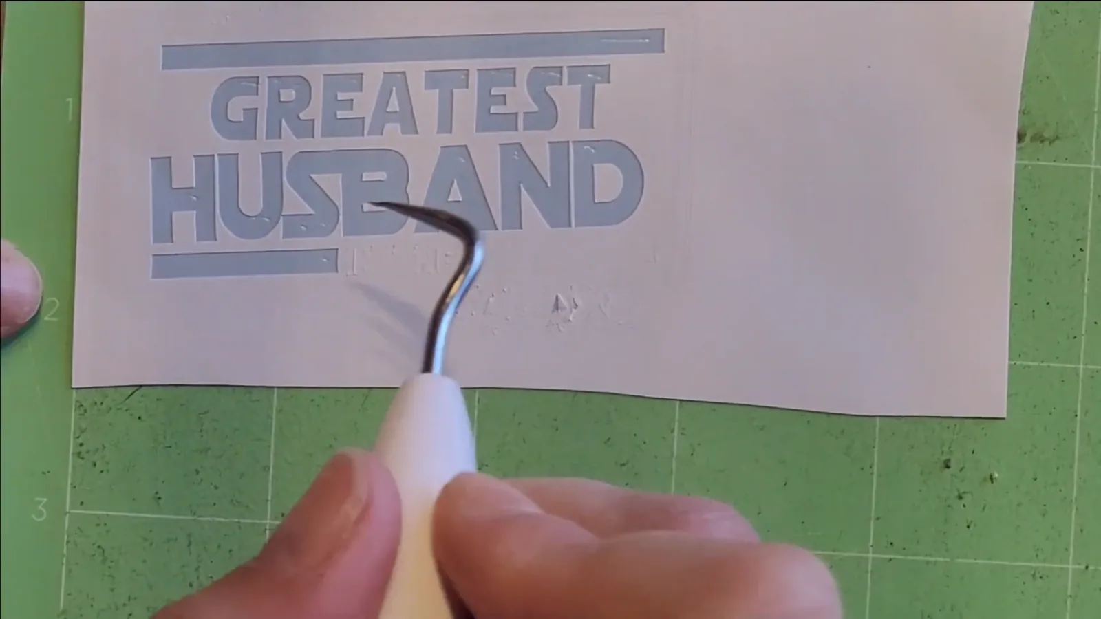 Weeding hook lifting letters out of a vinyl stencil on a green Cricut mat, leaving the surrounding vinyl in place for glass etching