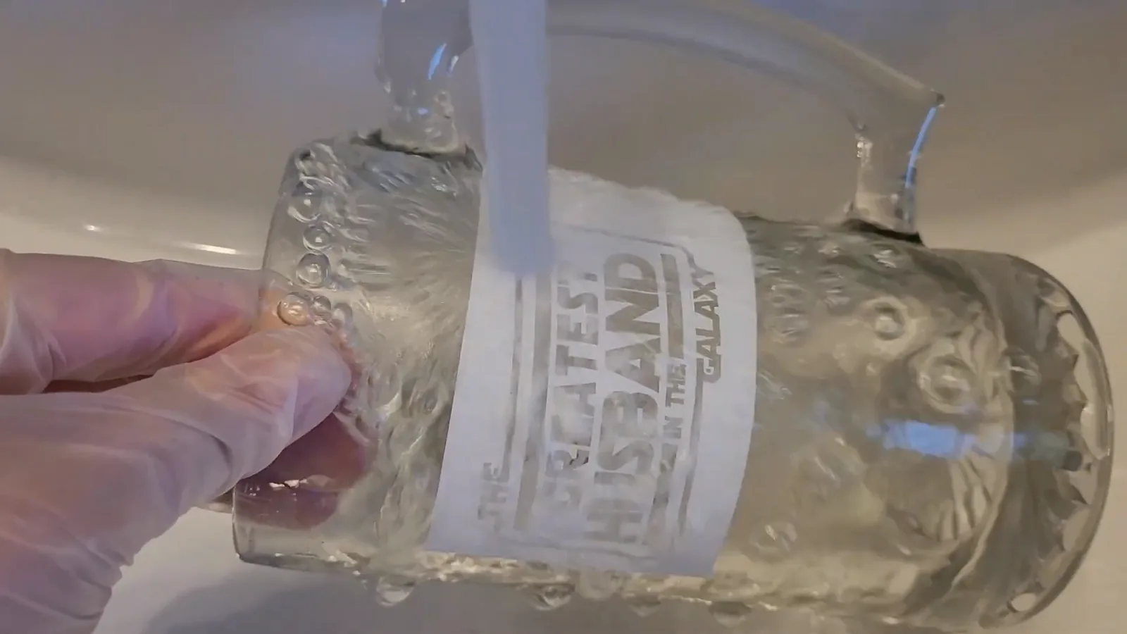 Gloved hand rinsing etching cream off a glass mug under cool running water in a sink, vinyl stencil still attached