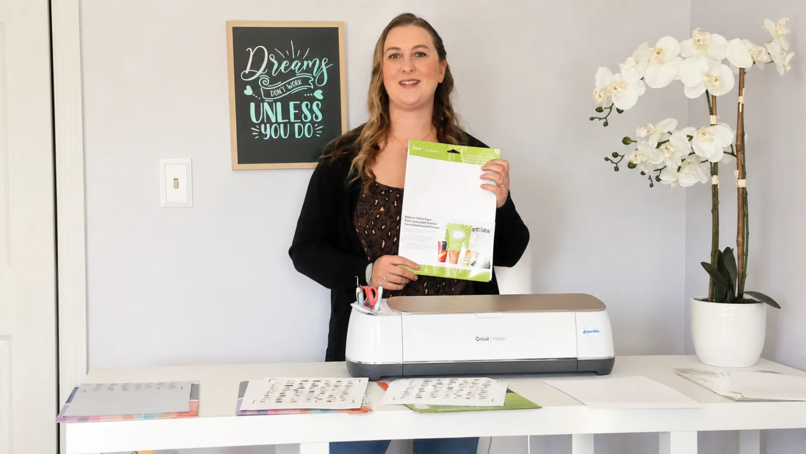 Kerri holding a package of Cricut Premium Printable Sticker Paper behind a Cricut Maker with printed sticker test sheets on the desk